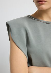 Primo piano di una persona che indossa un top grigio senza maniche con spalle strutturate e un colletto rifinito con strass.