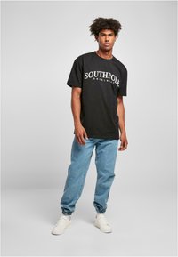 Juodas medvilnės marškinėliai su užrašu "SOUTHPOLE ORIGINALS" baltu spalva, derinami su šviesiai mėlynomis džinsinėmis kelnėmis ir baltais sportiniais batais.
