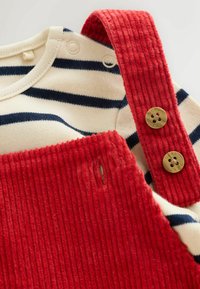 Rote Corduroy-Latzhose mit strukturierten Trägern und Messingknöpfen, kombiniert mit einem cremefarbenen Oberteil mit navyfarbenen horizontalen Streifen.