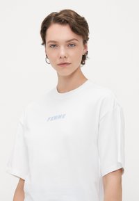 Vit bomull t-shirt med korta ärmar, med ljusblå "FEMME" text broderad på bröstet. Enkel, avslappnad passform.