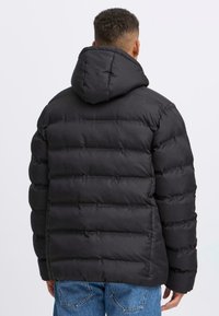 Mand i en sort quiltet pufferjakke med hætte, set fra siden, sammen med blå jeans foran en ensfarvet baggrund.