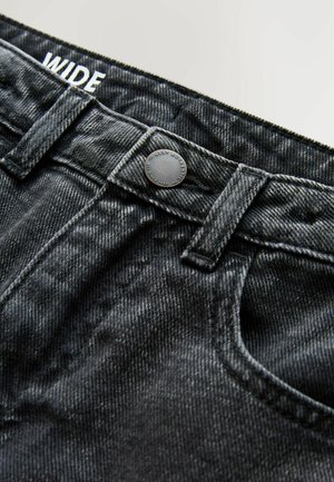 Jeans en denim noir large avec une surface texturée, dotés d'un bouton, d'une poche avant et de coutures visibles. L'étiquette indique "WIDE."