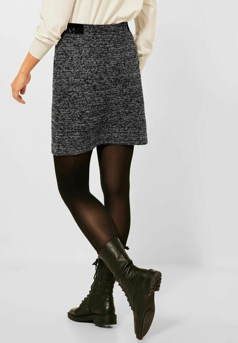 Femme portant une mini-jupe texturée noire et blanche, des collants noirs et des bottes de combat en cuir noir à lacets, debout, une jambe croisée derrière.