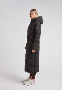 Abrigo largo negro tipo puffer con capucha, con un diseño acolchado, cremallera frontal y bolsillos laterales, combinado con zapatillas blancas y leggings marrones.