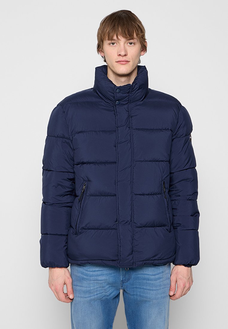Granatowa kurtka puchowa z wysokim kołnierzem, zamykanymi na zamek kieszeniami i pikowanym wzorem. Wykonana z gładkiego, syntetycznego materiału o luźnym kroju.