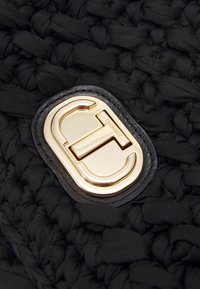 Borsa in tessuto intrecciato nero con chiusura a logo in tono oro. La texture è morbida con un motivo in rilievo e dettagli in pelle liscia attorno alla chiusura.