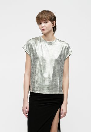 Mladić s kratkom kosom nosi metalik srebrnu majicu s kratkim rukavima i crnu suknju s visokim prorezom na plain pozadini.