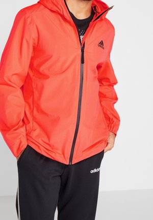 Red windbreaker jacket with a hood, featuring a black zipper and logo. Paired with black sweatpants displaying white branding. Smooth texture.