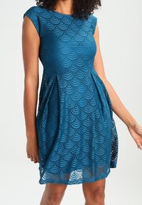 Robe en dentelle teal avec des manches courtes, motif festonné, silhouette évasée et taille froncée. Le tissu léger améliore la respirabilité.