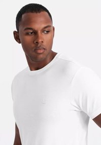 Biała koszulka z krótkim rękawem wykonana z gładkiego materiału, z okrągłym dekoltem i małym logo po lewej stronie klatki piersiowej.