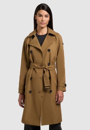 Žena s dlhými tmavými vlasmi, oblečená v zapnutom béžovom trenčkotu previazanom v páse, cez čierne tričko, stojaca pred jednoduchým svetlým pozadím.