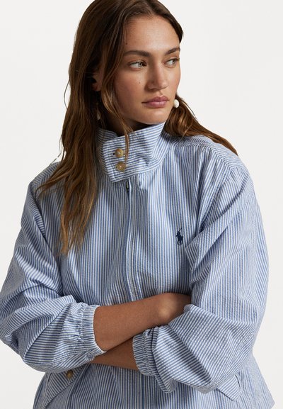 Casaco riscado em azul claro e branco, com gola alta, fecho de botão e punhos elásticos. Apresenta um pequeno logotipo em marinho no peito.