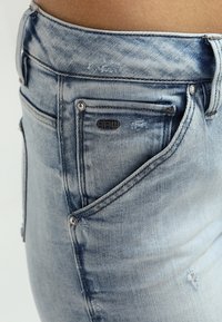 Jeans de mezclilla azul claro con acentos desgastados, un logo crudo cerca del bolsillo y un diseño entallado con detalles de costura visibles.