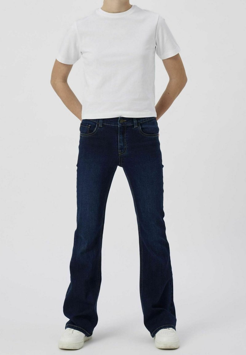 Personne portant un t-shirt blanc uni à manches courtes, un jean en denim bleu foncé et des baskets blanches, debout les mains dans le dos.