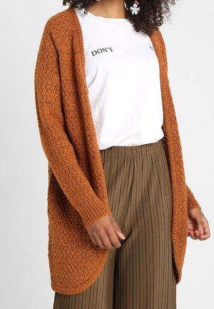 Femme portant un cardigan texturé orange brûlé sur une chemise blanche avec le texte "DON'T" et un pantalon large marron à fines rayures.
