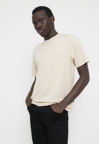 Μπεζ κοντομάνικη μπλούζα με στρογγυλή λαιμόκοψη και διακριτική λεπτομέρεια λογότυπου. Μαλακό ύφασμα, άνετη εφαρμογή, συνδυασμένη με μαύρο παντελόνι. Μινιμαλιστικός σχεδιασμός.