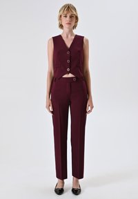 Completo composto da giacchino e pantaloni su misura color borgogna con un design corto, caratterizzato da grandi bottoni rotondi e una texture liscia. Scarpe slip-on nere lucide.