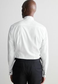Camicia bianca a maniche lunghe con colletto alto, con gemelli ai polsi, abbinata a pantaloni neri, vista da dietro su uno sfondo chiaro.