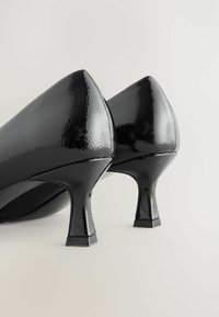 Scarpe con tacco alto in vernice nera lucida, con tacco quadrato, angolate leggermente per mettere in risalto la loro forma curva su uno sfondo neutro.