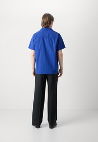 Camisa azul de manga curta com corte reto, apresentando colarinho e costura nas costas, combinada com calças largas pretas e sapatos pretos.