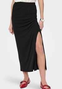 Zwarte maxi rok met een zijsplit, gemaakt van zachte stof, voorzien van een verzamelde taille en een gladde textuur. Gecombineerd met rode flats.