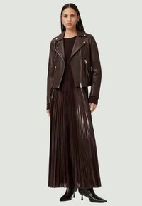 Jaqueta de motoqueiro de couro castanho sobre uma blusa escura, acompanhada por uma saia longa e plissada escura. Botas de salto alto pretas. Brincos simples visíveis.