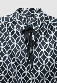 Schwarze Bluse mit einem geometrischen weißen Muster aus miteinander verbundenen Kreisen. Verfügt über einen Kragen und eine Schleife am Ausschnitt. Seidige Textur.
