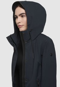 Marinblå hooded jacka med hel dragkedja, tryckknappar och justerbar dragsko. Har ett texturerat tyg och en logotyp patch på ärmen.