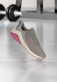 Grauer Sportschuh mit pinken Akzenten, Obermaterial aus Mesh, Schnürverschluss, Gummisohle und "AIR ZOOM"-Markierung an der Seite, vor einem Gym-Hintergrund.