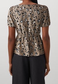 Donna che indossa una blouse corta a maniche corte con stampa leopardata beige e vita elastica, abbinata a pantaloni neri a gamba larga, vista da dietro.