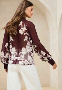 Sheer burgundy blouse with white floral pattern, features long puffed sleeves and buttoned cuffs. Smooth texture, elegant design.