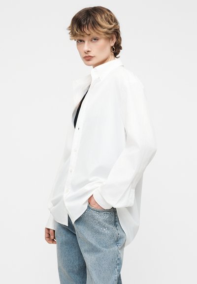 Άτομο με κοντά κυματιστά μαλλιά που φοράει άνετο λευκό πουκάμισο με κουμπιά πάνω από μαύρο τοπ, φωτεινό μπλε τζιν με διακριτικές διακοσμήσεις.