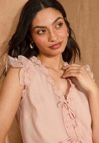 Blusa rosa pallido con maniche arricciate, scollo a V e fiocchetto decorativo. Tessuto morbido e leggero con una texture sottile e dettagli femminili.