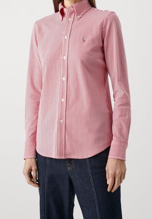 Kvinde iført en langærmet, rød- og hvidstribet skjorte med mørkeblå jeans, står mod en ensfarvet baggrund.