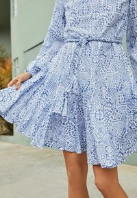 Robe bleu clair avec des motifs abstraits blancs, manches longues et ceinture à la taille ; dotée d'une jupe évasée avec des volants superposés.