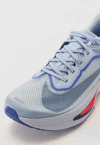 Hellblauer Sportschuh mit Mesh-Details, runder Form, lila Akzenten, auffälligem Logo und rotem Fersenbereich. Robuste Gummisohle.