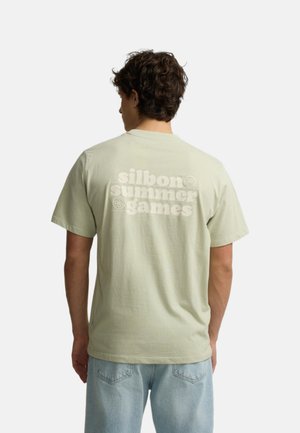 Joven con pelo rizado que lleva una camiseta verde claro con "silbon summer games" impreso en la parte trasera y vaqueros azul claro.