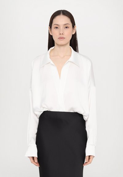 Bílá halenka s dlouhým rukávem, hlubokým výstřihem do V a oversized střihem, doplněná černou tužkovou sukní. Hladká tkanina, minimalistický design a manžetové rukávy.