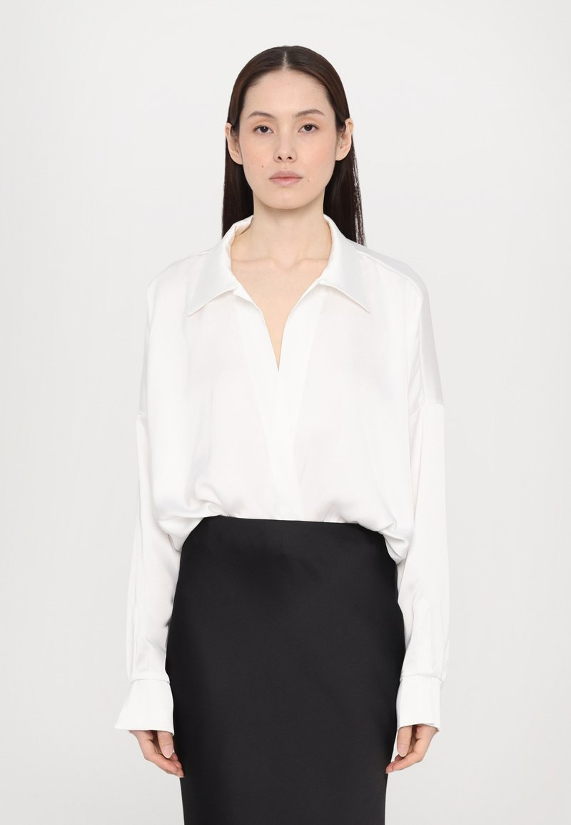 White long-sleeve blouse with a deep V-neck and oversized fit, paired with a black pencil skirt. Smooth fabric, minimal design, and cuffed sleeves.