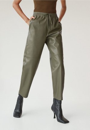 Mujer con pantalones deportivos de cuero verde oliva con cintura de cordón y botines negros de tacón, de pie con las manos en los bolsillos.