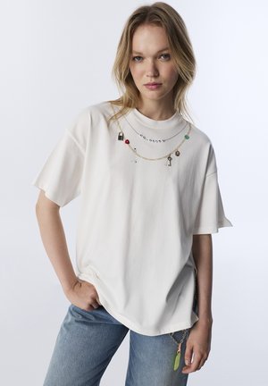 T-shirt blanc oversize à manches courtes, orné de colliers à charms superposés, avec une coupe décontractée et un pantalon en denim texturé.