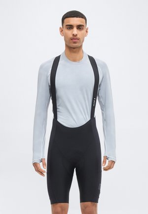 Férfi világosszürke hosszú ujjú felsőben és fekete kerékpáros kantáros rövidnadrágban, egyszerű háttér előtt állva.