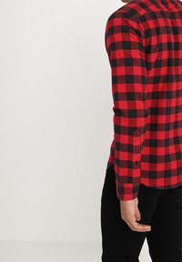 Camisa de franela a cuadros rojos y negros con mangas largas, puños con botones y cuello redondeado, llevada sobre pantalones negros.