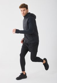 Melna polsterēta veste ar kapuci, kas uzvilkta virs gara piedurkņu tops, apvienota ar melniem šortiem un legingiem, kā arī melniem sporta apaviem.