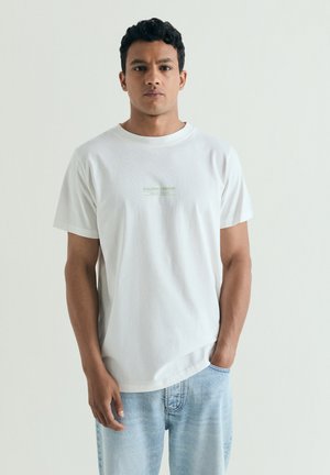Hombre con cabello corto y rizado, con camiseta blanca y vaqueros azul claro, de pie frente a un fondo liso y claro, con una mano en el bolsillo.