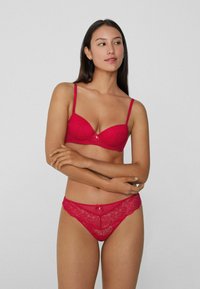 Červený krajkový prádlo set, který obsahuje vyztuženou podprsenku s nastavovacími ramínky a odpovídající tanga s malou mašlí a krajkovým lemem.