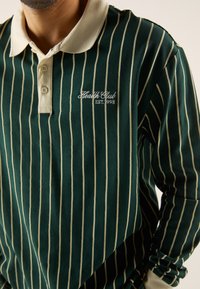 Groen gestreepte lange mouw polo met een crèmekleurige kraag. Bevat geborduurd detail "Health Club EST. 1998". Zachte, gestructureerde katoenstof.