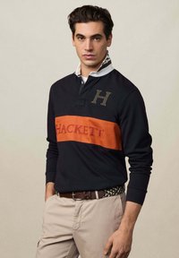 Mornarsko plava duga polo majica s narančastim prugama, logom "HACKETT" i rebrastim ovratnikom. Uparena sa bež hlačama i remenom s uzorkom.