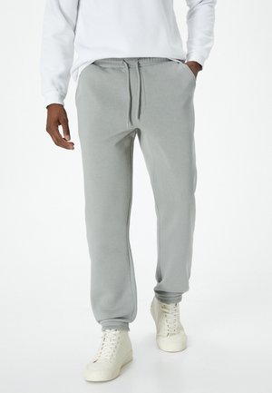 Pantalones de chándal grises con cintura elástica y cordón ajustable, con bolsillos laterales y puños acanalados, usados con zapatillas blancas.