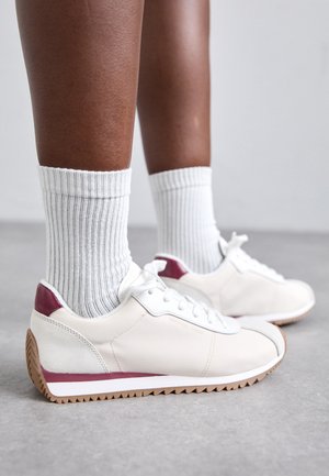 Sneaker bianchi con dettagli bordeaux, suole texturizzate, tomaia in pelle liscia e punta rotonda. Indossate con calze bianche a coste.